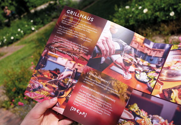 Abgebildet ist die Innenseite eines Flyers für Grillhaus. Er wird gehalten von einer Hand und es ist Natur im Hintergrund zu sehen.